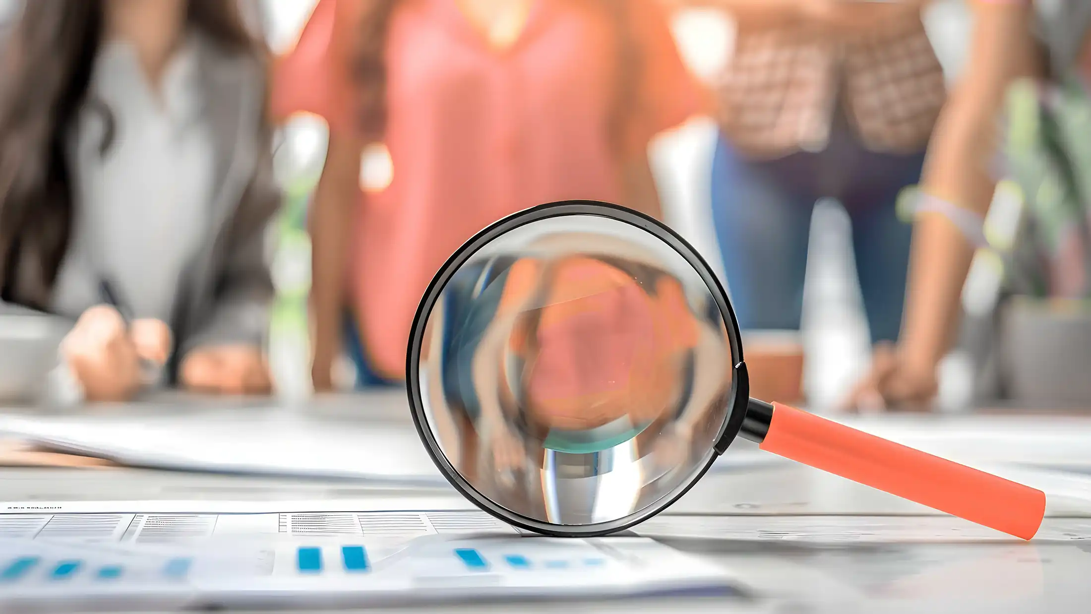A magnifying glass sites on a table in the foreground, blurred people are lined up in the background.