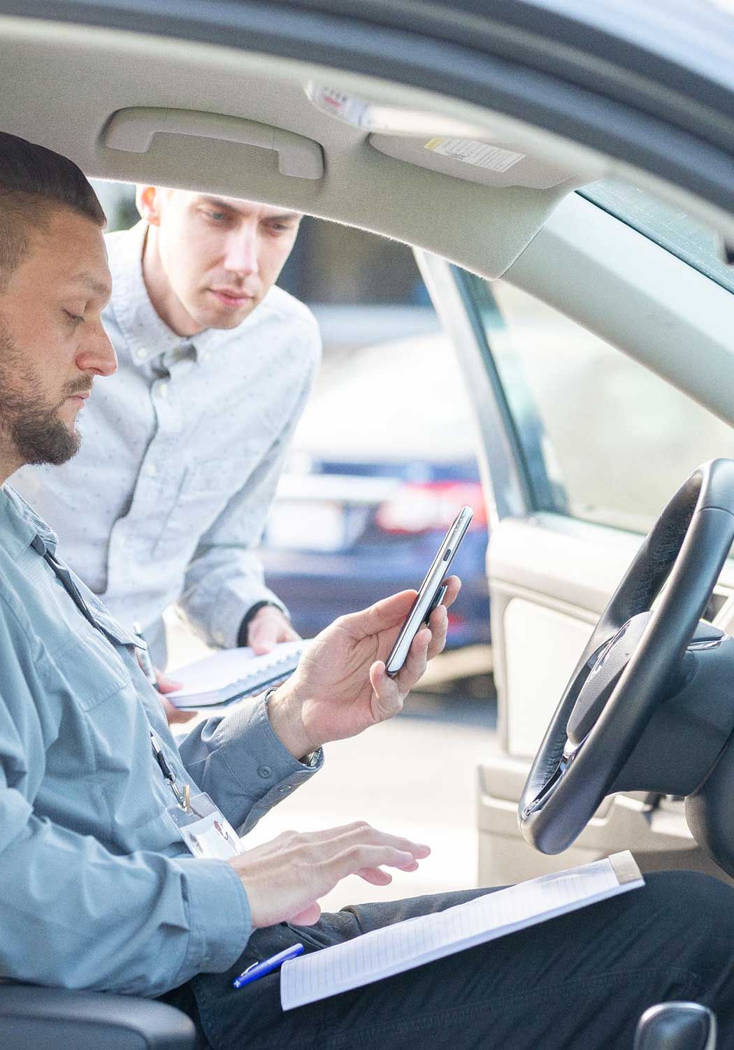 Employee in a vehicle at a dealership writing down the details of a car on a notepad and taking images on his phone.